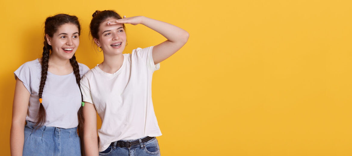 Happy smiling girl keeping palm near forehead and looking far away, standing against yellow wall, girl with dark hair wearing white t shirt. Copy space for advertisement.