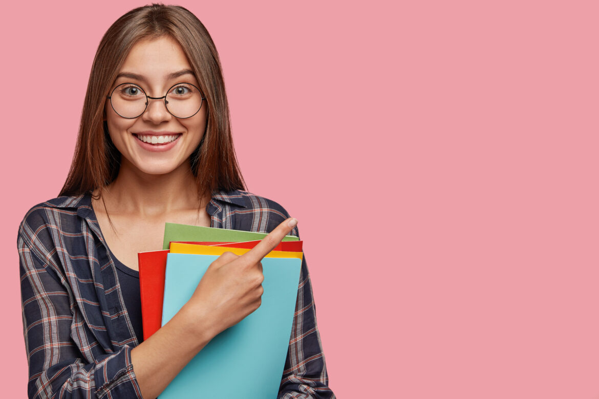 Studio shot of good looking young woman with dark hair, dressed in checkered shirt, points with index finger at copy space, holds books, advertises her university or college poses over pink background